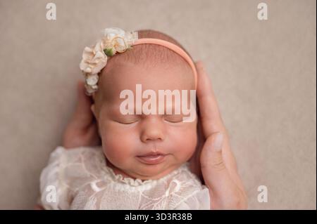 Un bébé nouveau-né serein et adorable portant un délicat bandeau floral dort paisiblement, bercé dans de douces mains. Sa douceur évoque la chaleur Banque D'Images