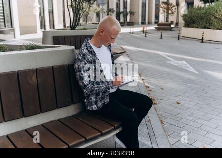 Un jeune homme à l'apparence albinos est assis sur un banc en bois dans une zone urbaine, écrivant des notes dans un cahier à reliure en spirale. Le cadre est moderne, avec s Banque D'Images