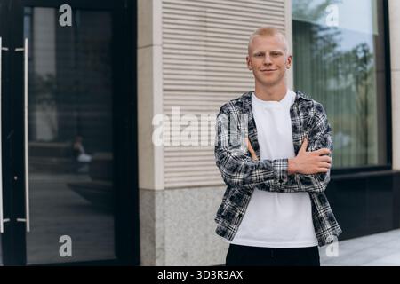 Un jeune homme à l'apparence albinos se tient confiant, les bras croisés, portant une chemise à carreaux décontractée et un tee-shirt blanc, devant un bâtiment moderne Banque D'Images