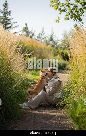 Une femme est assise sur un chemin, embrassant son chien dans un parc luxuriant pendant une journée ensoleillée, avec de grandes herbes qui les entourent, capturant un moment de pure joie et com Banque D'Images