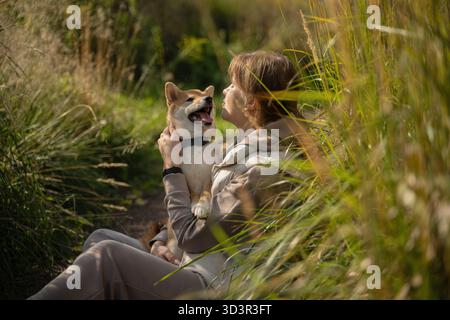 Une femme tient amoureusement son chien tout en se relaxant dans un champ ensoleillé pendant une promenade. Le moment serein souligne le lien entre animal et propriétaire au milieu de Lush Banque D'Images
