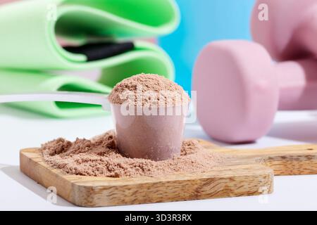 Une image en gros plan de poudre de protéines de chocolat, reposant sur une planche de bois. En arrière-plan, des haltères roses et un tapis vert enroulé suggèrent une séance d'entraînement Banque D'Images