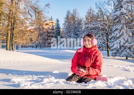 Une femme joyeuse dans un manteau rouge et des cache-oreilles roses profitant de la neige d'hiver dans les Alpes comme le soleil se couche, créant une lueur chaude sur le paysage enneigé A. Banque D'Images