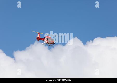 Un hélicoptère rouge éclatant survole des nuages blancs moelleux contre un ciel bleu vif dans les montagnes suisses près de Zermatt, capturant l'essence de l'Avent Banque D'Images