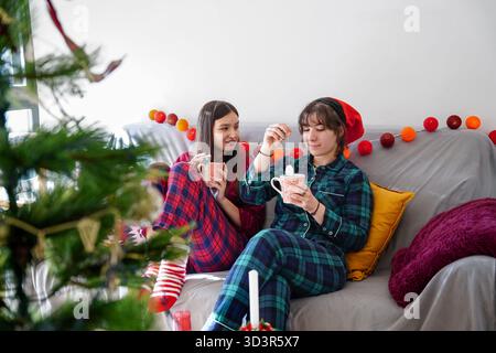 Deux filles en pyjama festif apprécient le chocolat chaud tout en se relaxant sur le canapé. La chambre décorée dispose d'un sapin de Noël et de lumières chaudes, créant un cadre confortable Banque D'Images