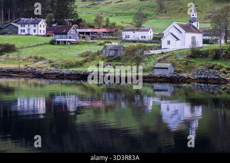 Église en bois blanc, hangar à bateaux et maisons reflétés dans un fjord calme près d'une colline boisée à Vestland, Norvège Banque D'Images