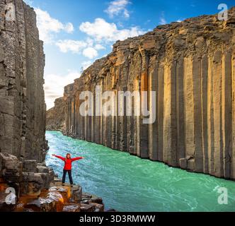 Une personne se tient debout les bras tendus sur un terrain rocheux dans le canyon de Studlagil, en Islande. Les colonnes de basalte hexagonales frappantes encadrent un turquoise éclatant Banque D'Images