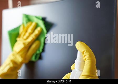 Gros plan des mains portant des gants jaunes nettoyant un moniteur avec un chiffon vert et un vaporisateur. Banque D'Images