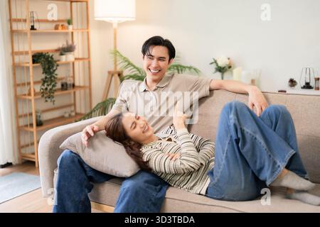 Jeune femme homme asiatique appréciant la vie à la maison confortable sur le canapé. Couple liant rire passer du temps de qualité ensemble. Atmosphère intime et détendue. Banque D'Images