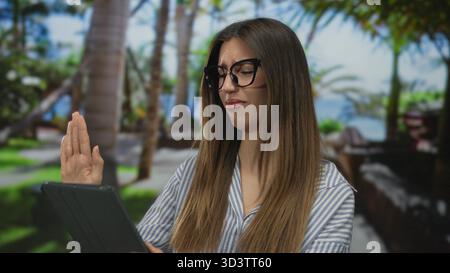 Femme tenant la tablette avec la main levée dans le parc vert parmi les palmiers portant une chemise rayée ; rejet dégoût aversion. Banque D'Images