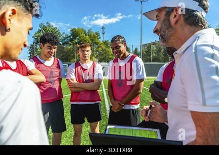 Entraîneur de soccer discutant de stratégie de jeu et expliquant les tactiques sur une tablette numérique avec un groupe de jeunes joueurs masculins portant des bavoirs de sport pendant l'entraînement Banque D'Images
