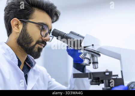 Scientifique aux cheveux foncés mène des expériences, ajuste le spécimen dans une boîte de Pétri et le regarde au microscope, faisant des recherches dans un laboratoire moderne Banque D'Images
