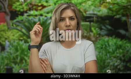 Femme portant un t-shirt blanc pointe l'index vers le haut dans une clairière forestière sous la canopée verte ; curiosité. Banque D'Images
