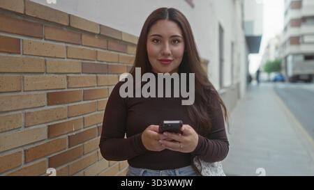 Femme hispanique fait défiler le smartphone sur la rue de la ville par le mur de briques tout en lisant des messages ; contentement calme. Banque D'Images
