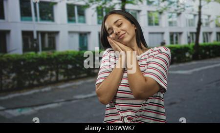 Femme pressant les mains sur la joue avec les yeux fermés sur une rue urbaine près d'un bâtiment, tshirt rayé noué, pose détendue et sourire subtil ; sérénité daydr Banque D'Images