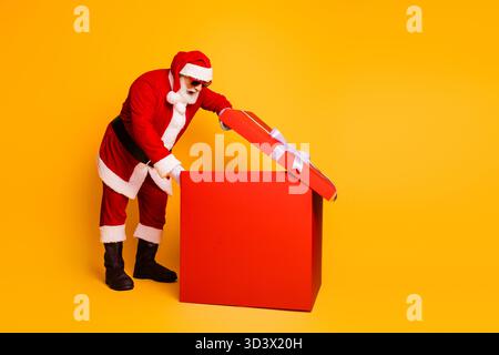 Santa ouvre une grande boîte cadeau rouge dans un studio jaune vif pour un joyeux moment de noël de cadeau de vacances et de joie Banque D'Images