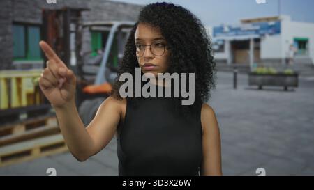 Femme portant des lunettes se tient à l'extérieur dans une zone industrielle avec un chariot élévateur et des bâtiments en arrière-plan, regardant concentré et pointant. Banque D'Images