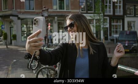 Femme prenant selfie et souriant avec smartphone à amsterdam, marchant le long de canaux pittoresques dans une tenue élégante pendant une journée ensoleillée mettant en vedette l'emblématique c hollandais Banque D'Images