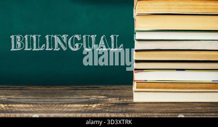 Pile de divers livres colorés reposant sur une table en bois rustique, avec un fond de tableau noir affichant le mot bilingue, créant un enseignement Banque D'Images