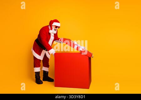 Santa dans un costume rouge ouvre une grande boîte cadeau sur un fond jaune vif pour les achats de Noël et la promotion des vacances Banque D'Images