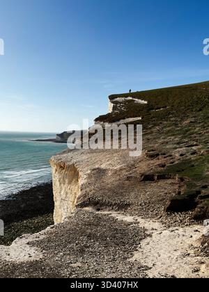 Scène de falaise côtière à Seven Sisters Cliffs montrant la mer bleue, le chemin de galets, et un marcheur lointain le long du bord. Banque D'Images