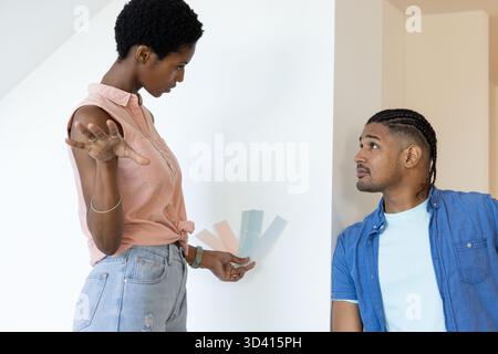 Couple diversifié sélectionnant des échantillons de peinture dans une pièce intérieure lumineuse, tenant des cartes de couleur pastel Banque D'Images