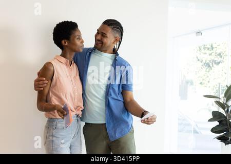 Tenant des échantillons de peinture et des cartes d'échantillon, couple diversifié choisissant des couleurs de décor dans une maison moderne lumineuse Banque D'Images