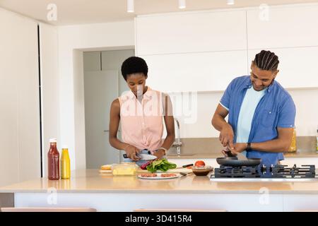 Cuisiner divers couples tranchant des légumes verts feuillus et remuant une poêle à l'îlot de cuisine moderne, avec du fromage Banque D'Images