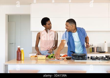 Couple diversifié hachant la laitue et arrangeant les petits pains au comptoir de cuisine, avec des condiments Banque D'Images