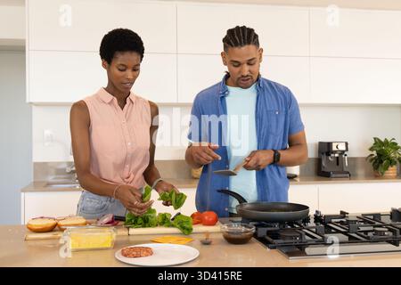 Cuisine de couple diversifiée ensemble à l'îlot de cuisine, avec des ingrédients de hamburger et une spatule Banque D'Images