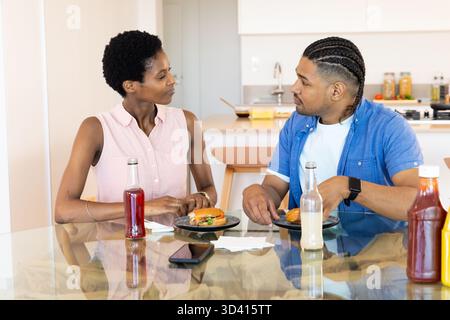 Couple diversifié mangeant des hamburgers à la table à manger en verre dans la cuisine, avec des bouteilles de soda et ketchup Banque D'Images