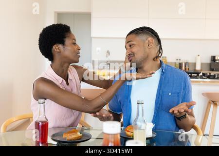 Geste couple diversifié bavardant à la table en verre dans la cuisine moderne, avec des sandwichs et des bouteilles Banque D'Images