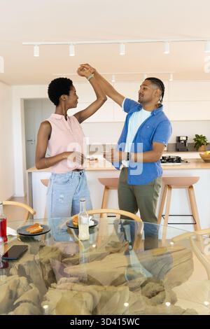 Couple de danse varié filant près de la table ronde avec plateau en verre dans la cuisine moderne, avec des articles pour le petit déjeuner Banque D'Images