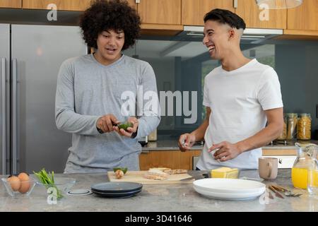 Divers amis masculins coupant l'avocat et craquant l'œuf dans la cuisine à la maison, avec du pain tranché et du jus Banque D'Images