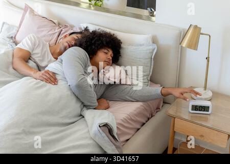Couché divers partenaires masculins étirant le bras vers le réveil numérique par table de nuit dans la chambre Banque D'Images