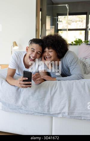 Rire divers amis masculins couchés sur le lit dans la chambre avec couette et oreillers gris, avec smartphone Banque D'Images