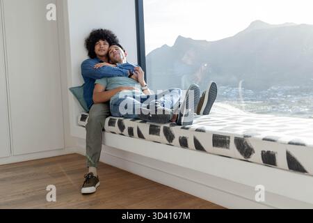 Divers partenaires masculins câlins dans un banc de fenêtre moderne, montrant coussin à motifs et vue sur la montagne Banque D'Images