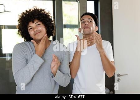 Divers amis masculins appliquant mousse à raser et nettoyant dans la salle de bain, avec miroir et panneaux de douche Banque D'Images