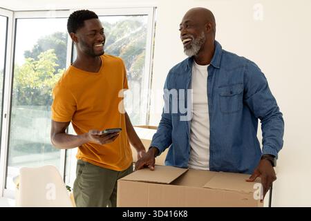 Sourire père afro-américain et fils déballant la boîte de déménagement à la maison, avec smartphone, chaise blanche Banque D'Images