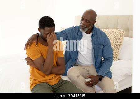 Réconfortant père afro-américain et fils assis sur le lit dans une chambre lumineuse, avec tête de lit touffetée Banque D'Images