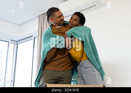 Partage couverture de jet sarcelle, couple diversifié debout à la maison près des fenêtres coulissantes, avec boîte mobile Banque D'Images