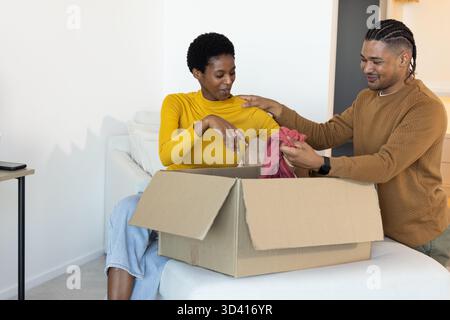 Déballage du couple diversifié assis sur le canapé dans le salon, avec boîte en carton et smartphone Banque D'Images