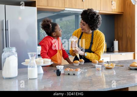 Tuyauterie diversifiée mère et fille faisant des cupcakes à l'île de marbre, avec des saupoudres Banque D'Images