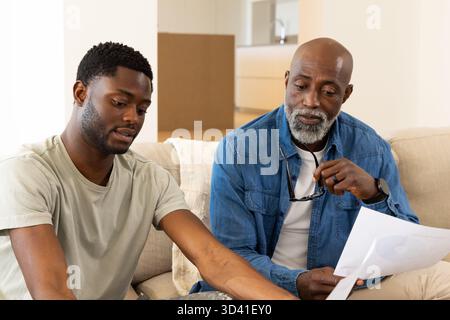 Père afro-américain et fils examinant les documents imprimés sur le canapé à la maison avec la boîte de déménagement Banque D'Images