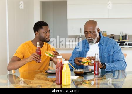 Père et fils afro-américains mangeant des hamburgers à table vitrée dans la cuisine, avec soda et ketchup Banque D'Images