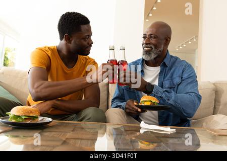 Clinking père afro-américain et fils partageant des bouteilles sur le canapé à la maison, avec des hamburgers sur la table Banque D'Images