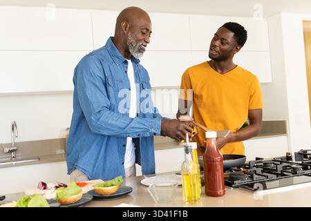 Père aîné afro-américain et fils cuisinant dans une cuisine moderne, avec spatule et petits pains à hamburger Banque D'Images