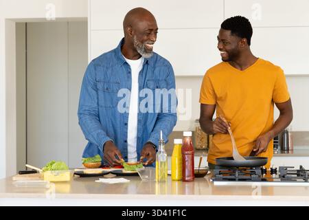 Souriant père afro-américain et fils préparant le repas à l'îlot de cuisine, avec spatule et bols Banque D'Images