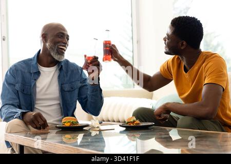 Clinking père afro-américain et fils assis sur le canapé dans le salon, avec thé glacé, hamburgers Banque D'Images