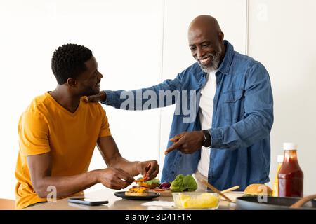 Père et fils afro-américains préparant des hamburgers dans une cuisine moderne, avec petits pains et smartphone Banque D'Images